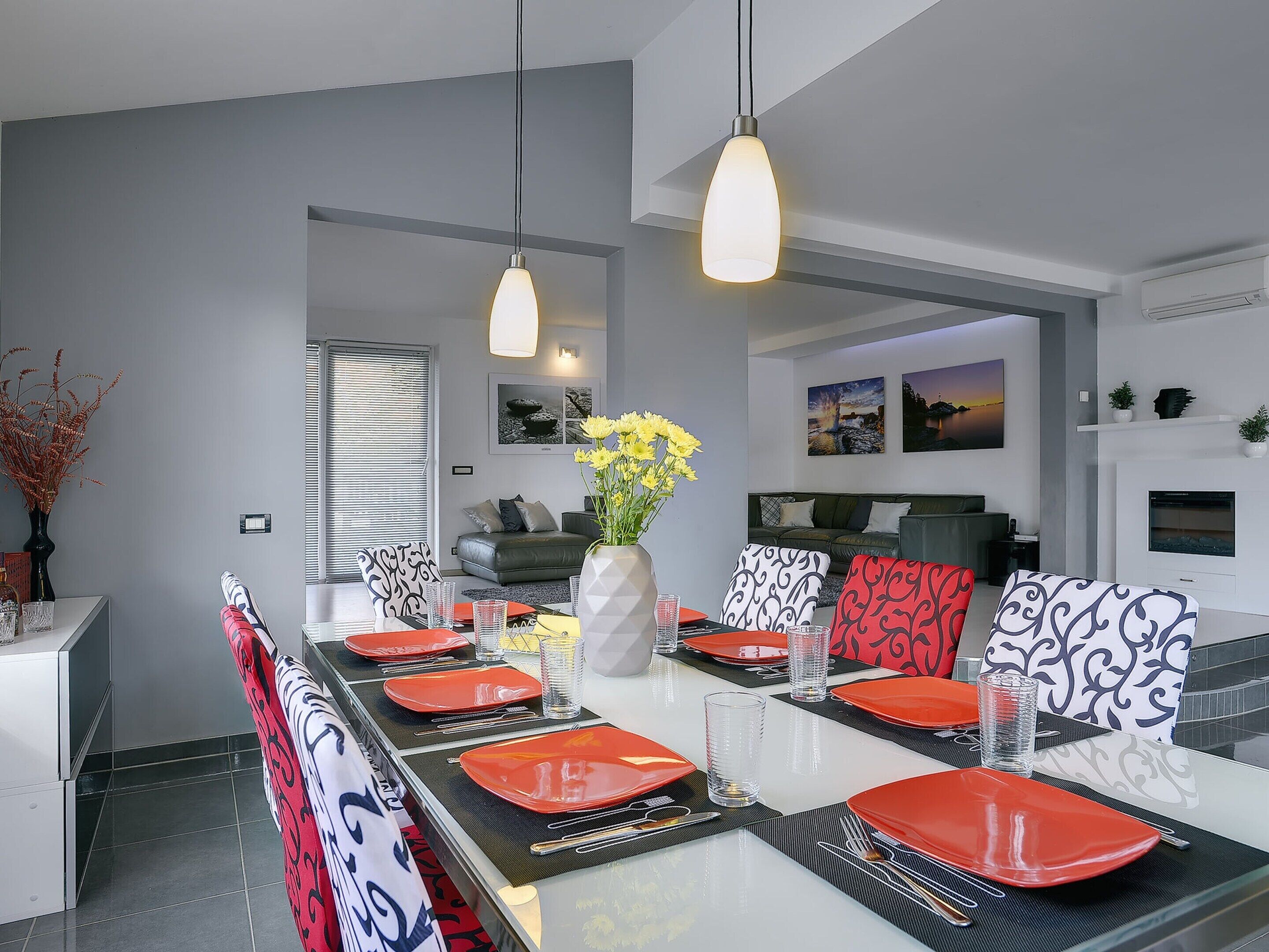 A modern dining room with a long table set for six, featuring red and black patterned chairs, a white vase with yellow flowers, and pendant lighting.