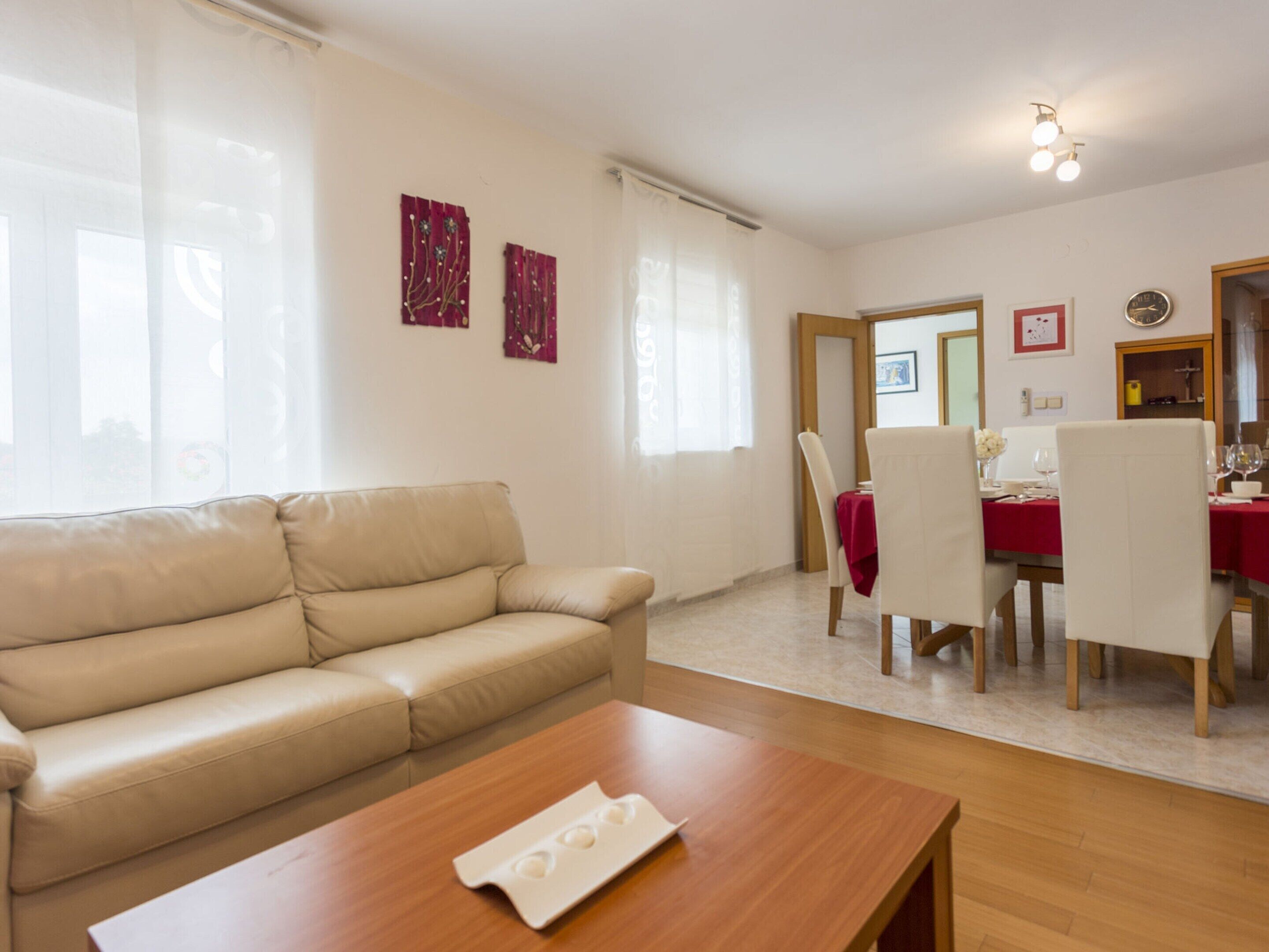 A living room with a beige sofa, a wooden coffee table, and a dining area with a red tablecloth and white chairs.