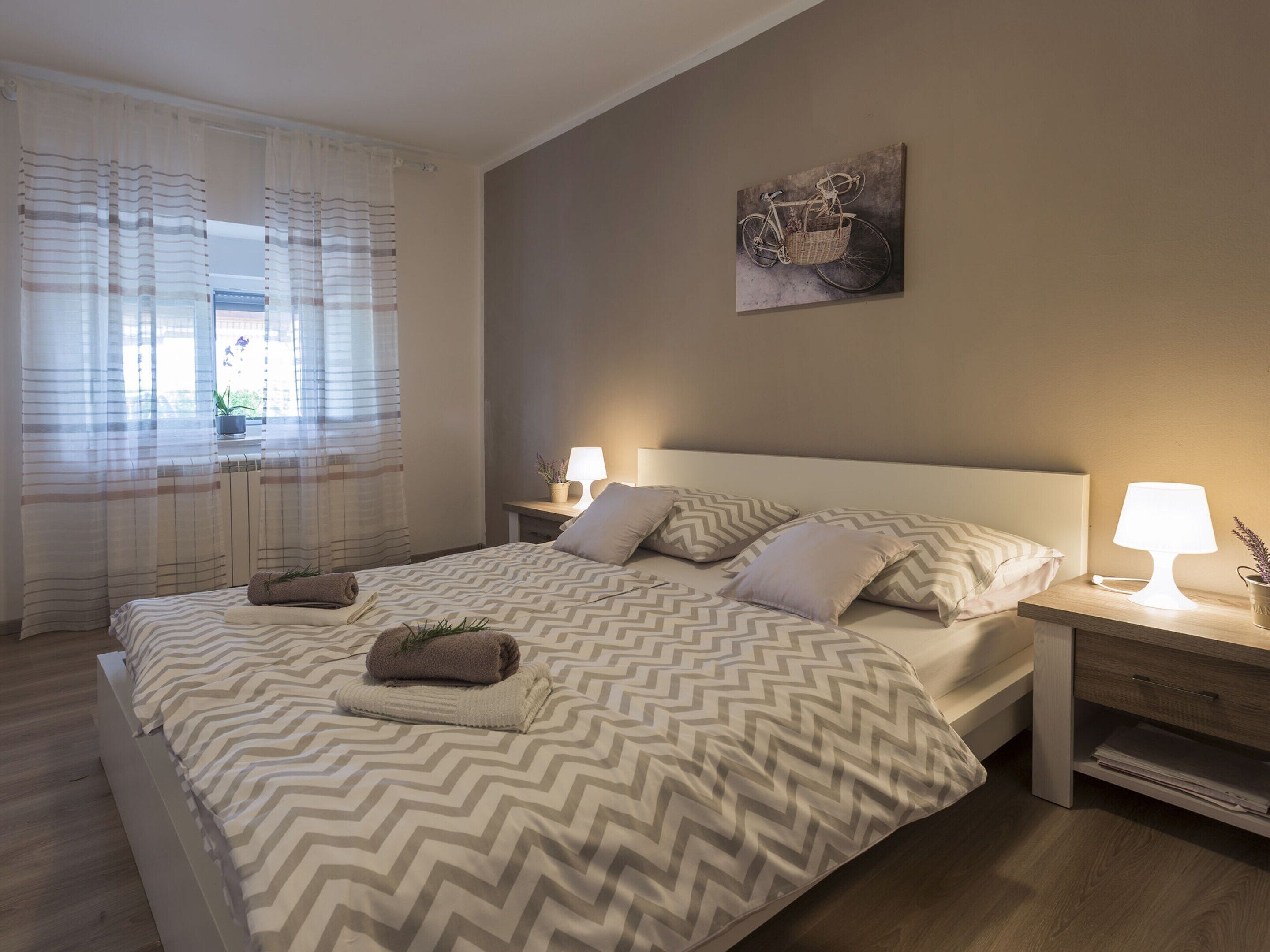 Un lit bien fait, recouvert d’une couvre-lit à motifs, avec deux lampes de chevet et un tableau représentant un vélo accroché au mur.