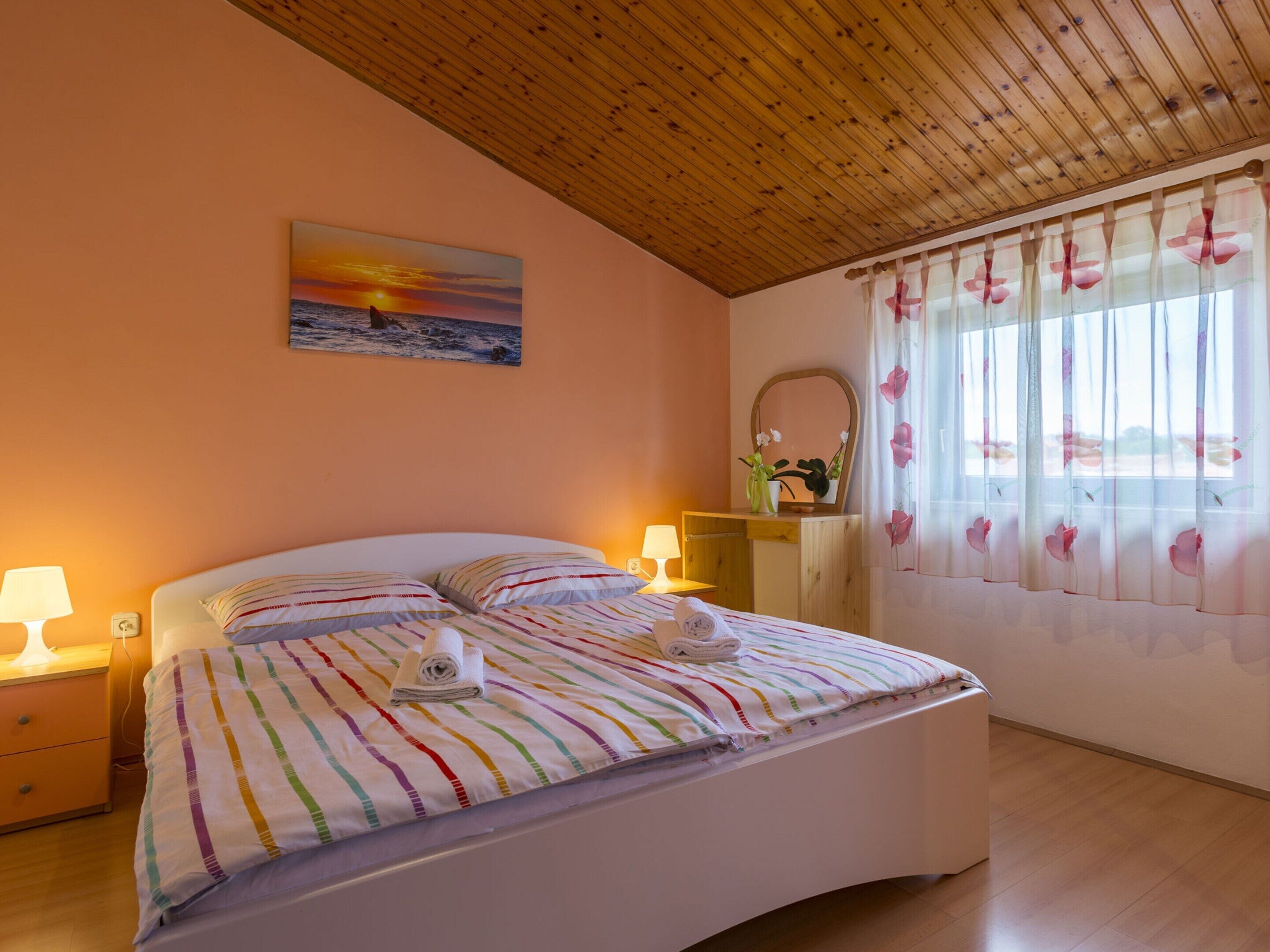Een slaapkamer met een houten plafond, een bed met gestreept beddengoed, een nachtkastje met een lamp, een dressoir met een spiegel, en een raam met bloemen gordijnen.
