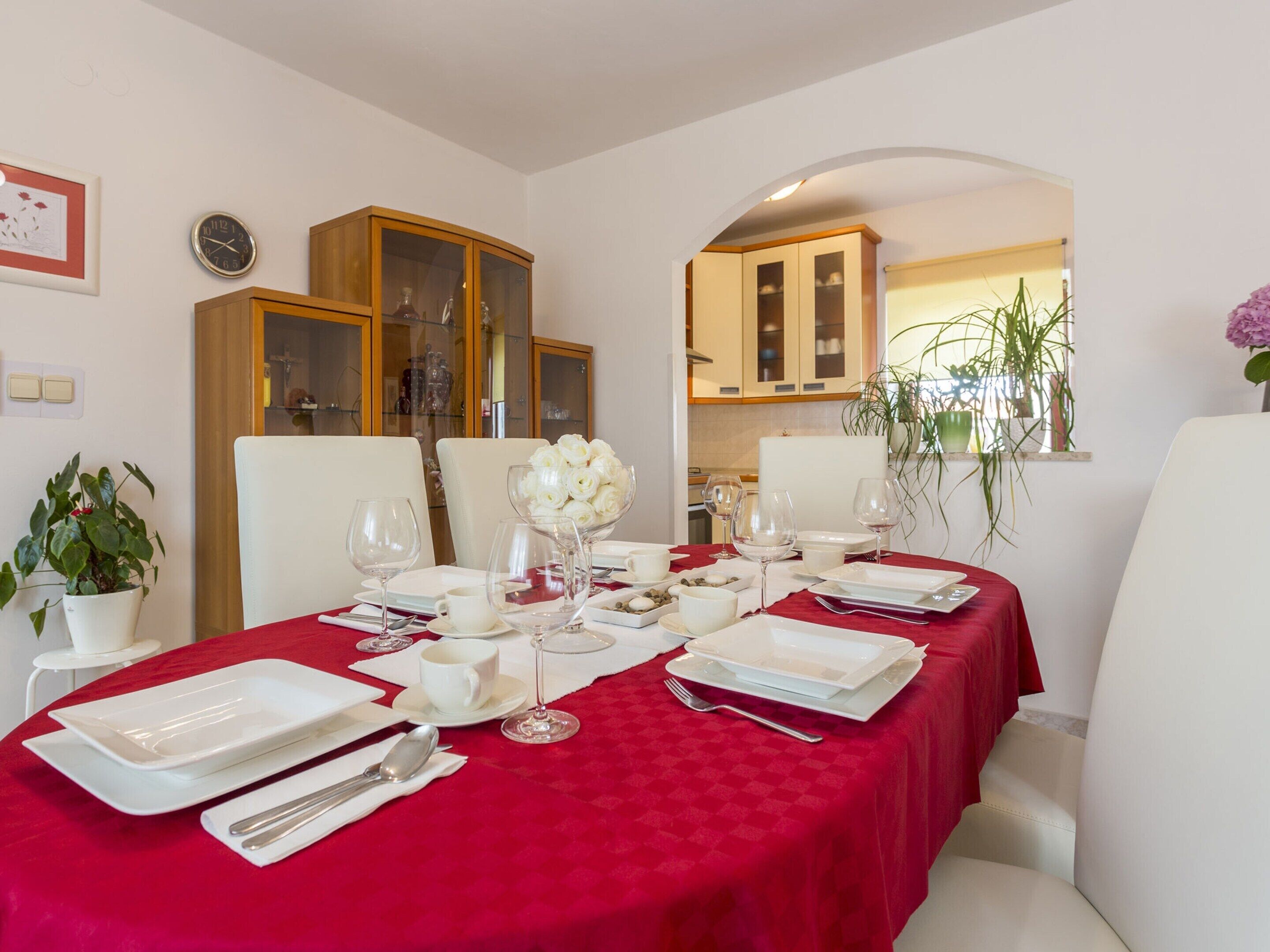 A dining area with a red tablecloth, white chairs, and a glass cabinet in the background.