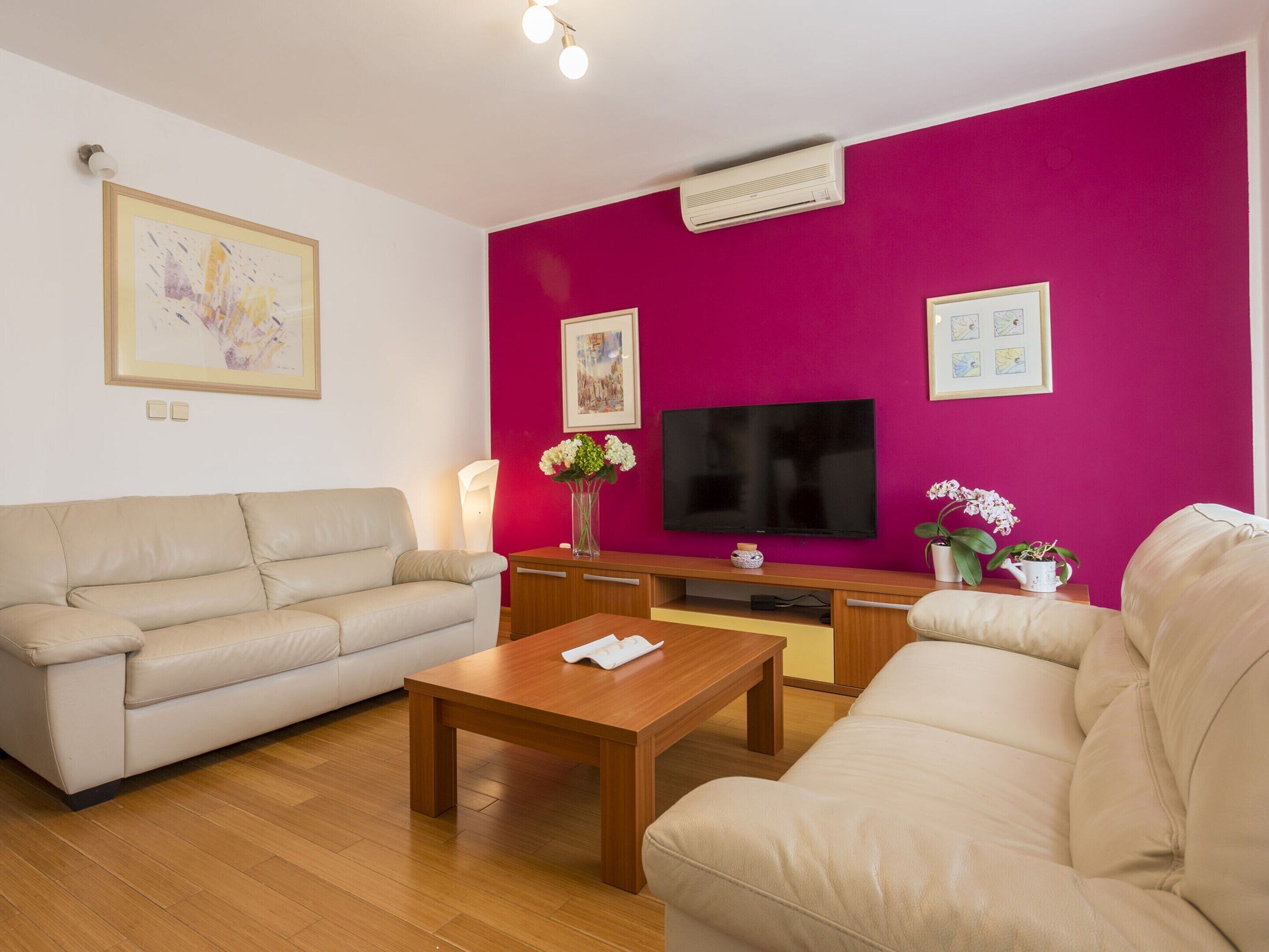 Een woonkamer met een beige bank, een houten salontafel, een flatscreen televisie en een roze accentmuur.