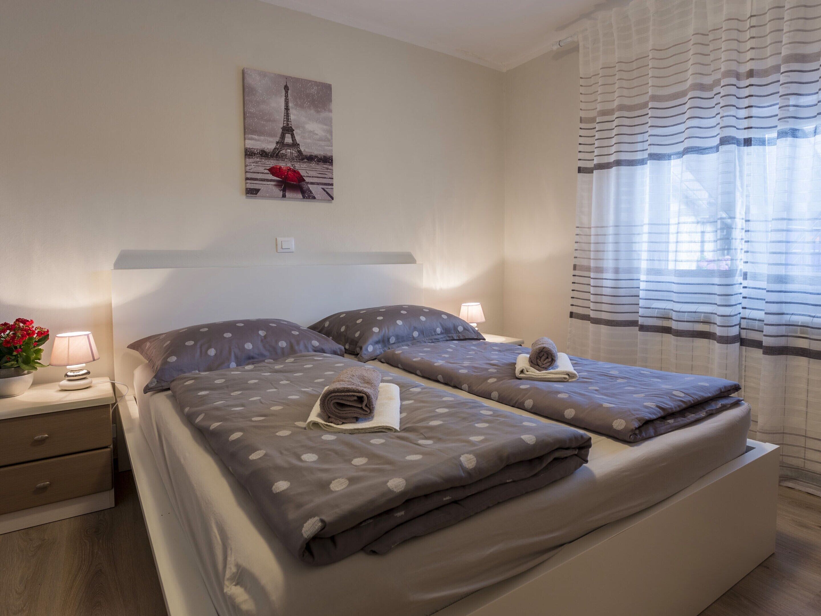 Habitación de hotel con dos camas, una mesita de noche con lámpara, un cuadro de la Torre Eiffel y una ventana con cortinas a rayas.