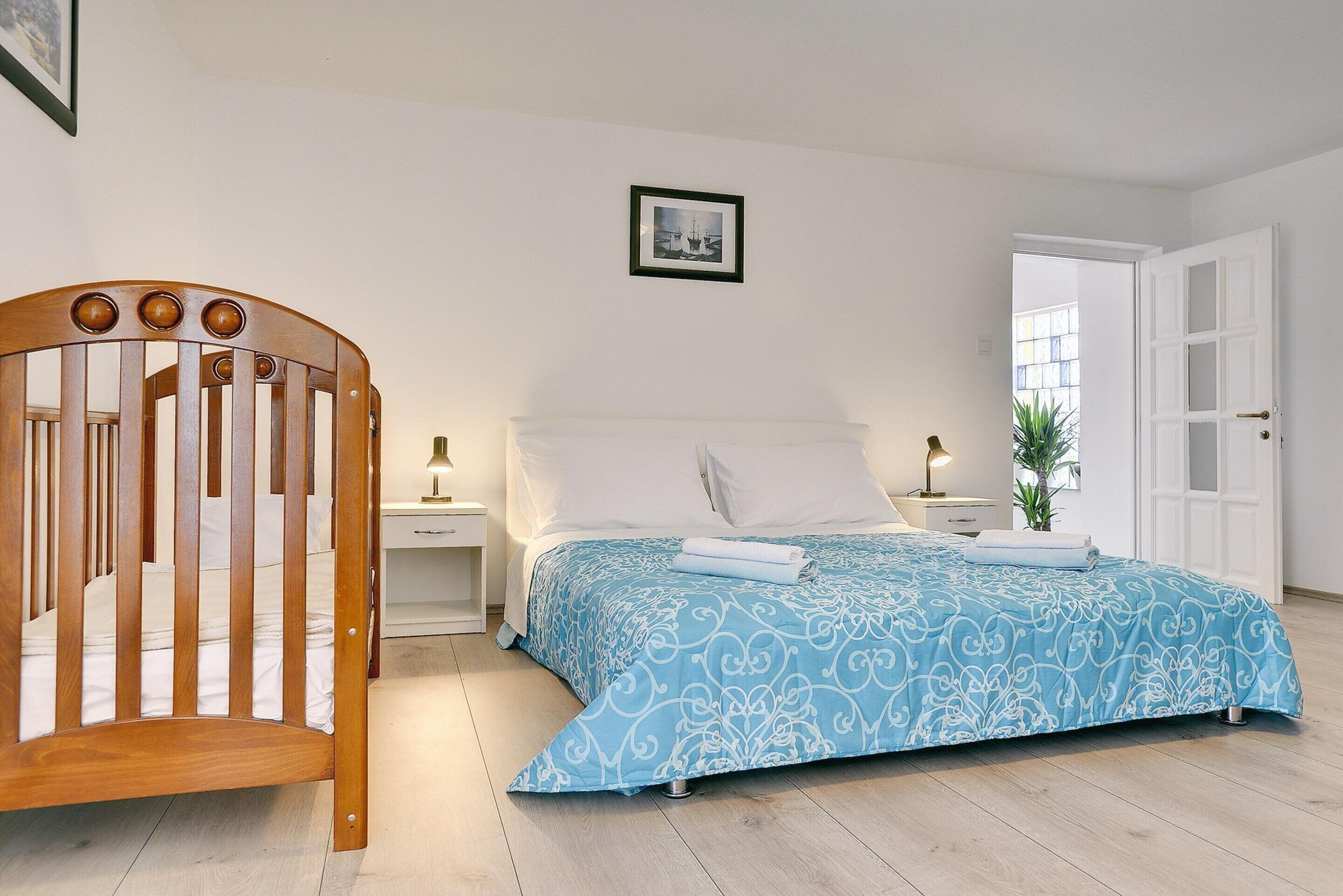 A bedroom with a wooden crib, a bed with a blue patterned bedspread, white pillows, and a framed picture on the wall.