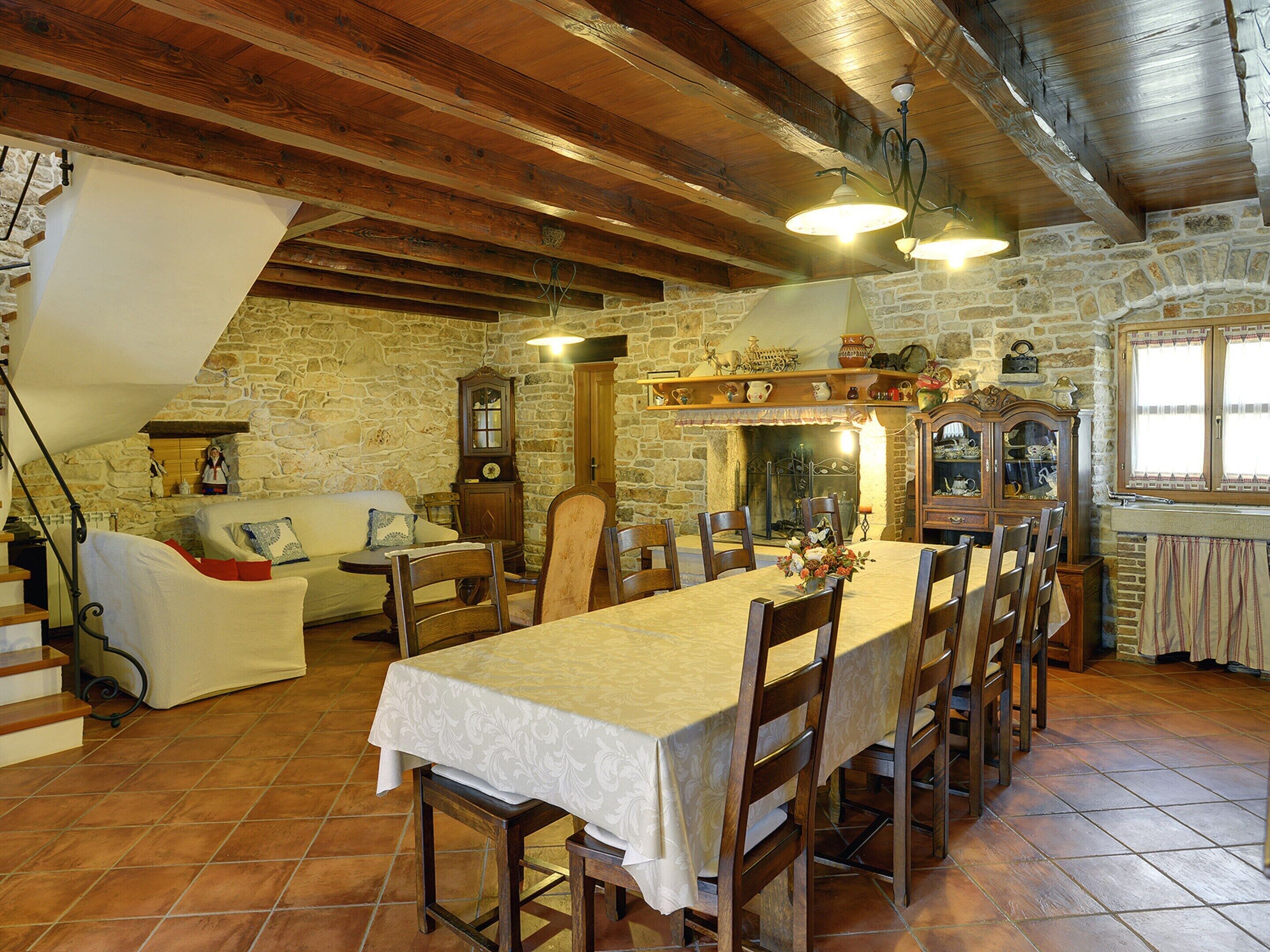 Un comedor con una mesa larga, sillas, techo de madera, paredes de piedra, chimenea y un reloj.