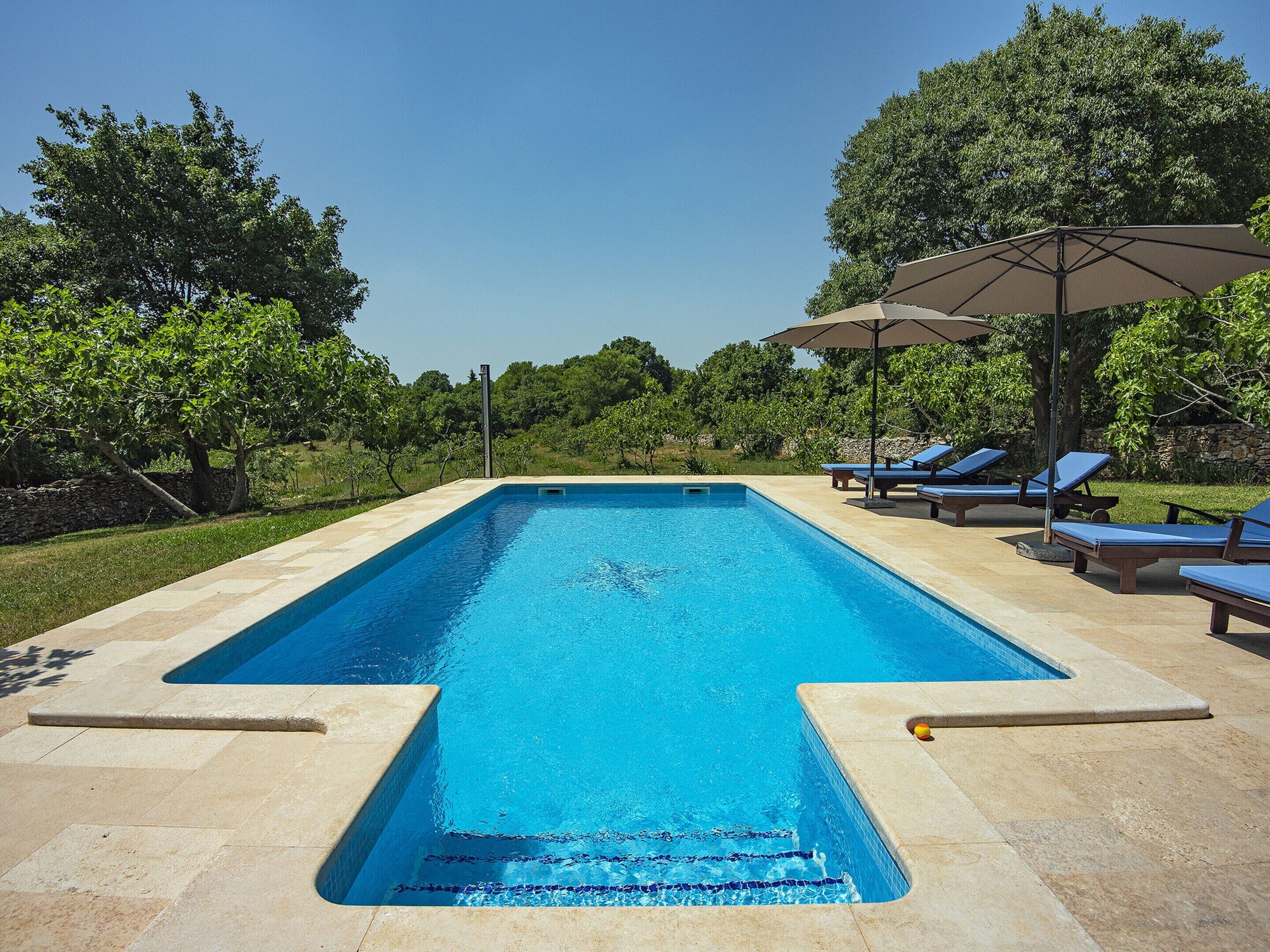 Ein rechteckiger Pool mit klarem, blauem Wasser, umgeben von einer Steindeck. Liegestühle und ein großer Sonnenschirm bieten Schatten.
