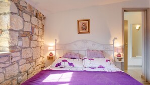 Un dormitorio con pared de piedra, una cama con ropa de cama de flores moradas, dos lámparas de noche y un cuadro en la pared.