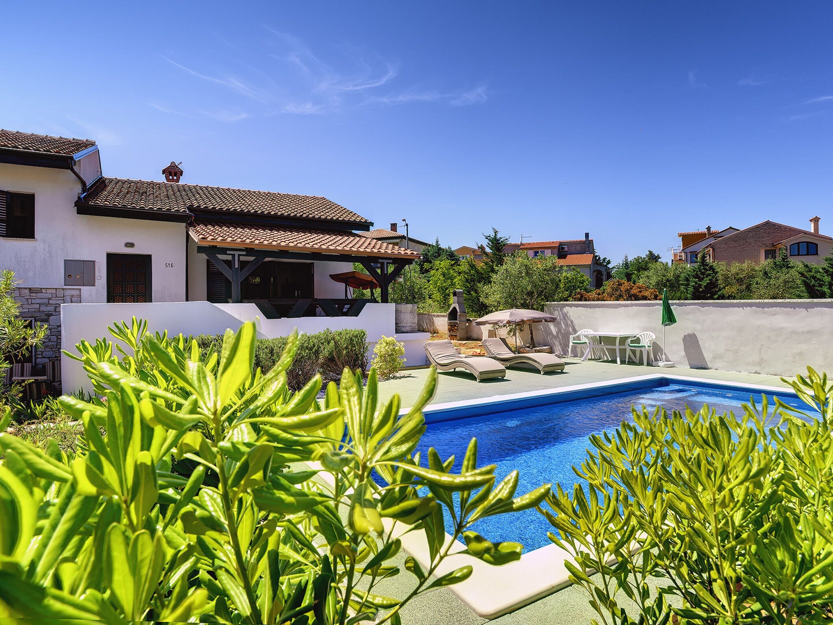 A pool area with lounge chairs and a barbecue, surrounded by greenery and a white building with a tiled roof.