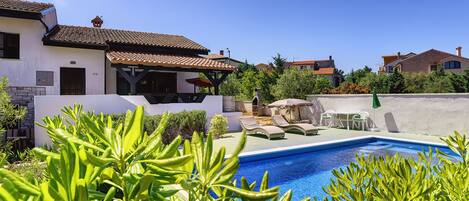 A pool area with lounge chairs and a barbecue, surrounded by greenery and a white building with a tiled roof.