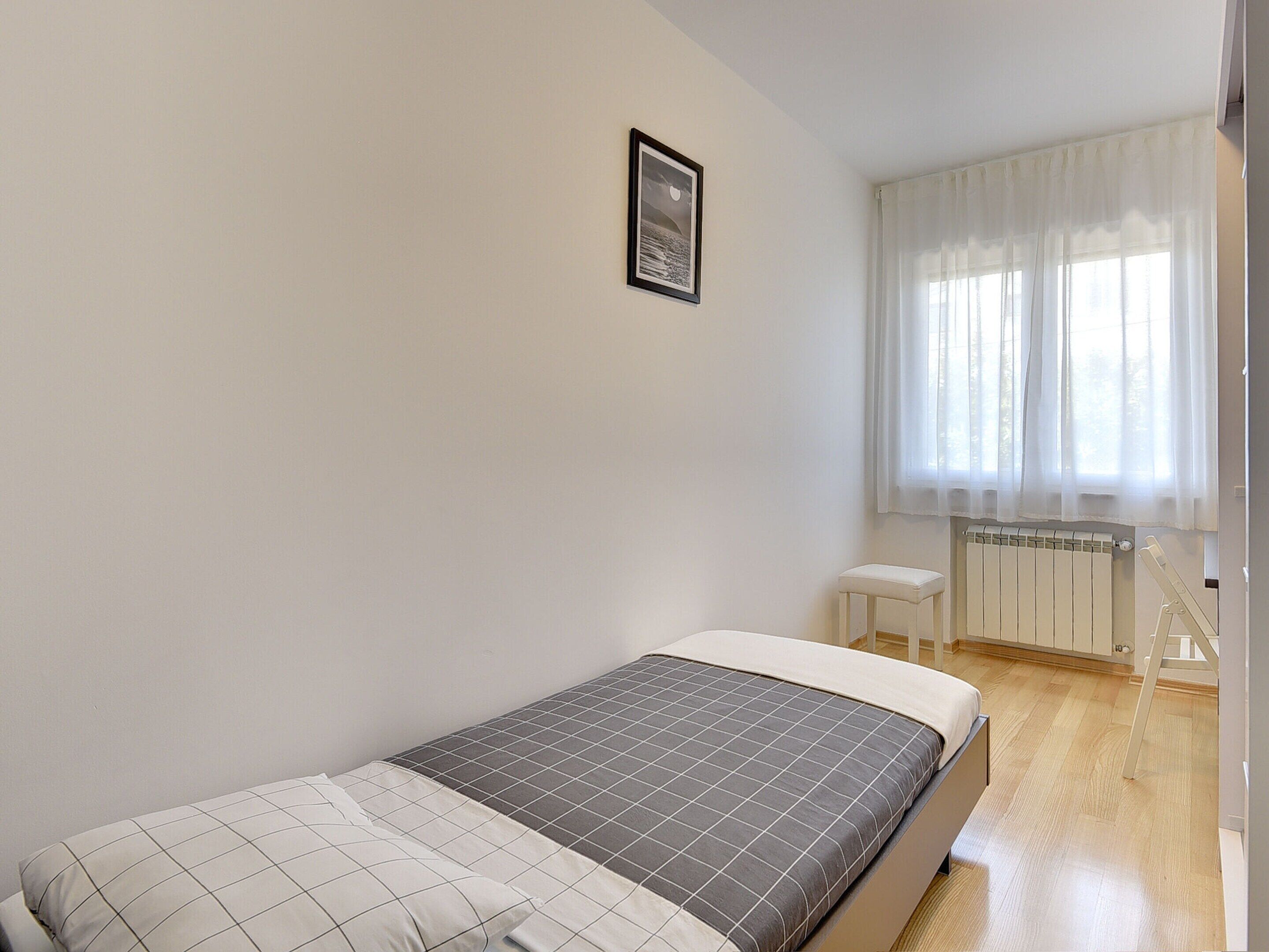 A bedroom with a bed, a small stool, a radiator, and a window with sheer curtains.