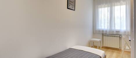 A bedroom with a bed, a small stool, a radiator, and a window with sheer curtains.