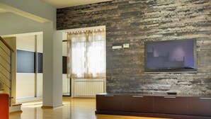 A modern living room with a flat-screen TV mounted on a textured wall, wooden flooring, and a glass coffee table. - Modern Holiday Home With Swimming Pool, Terrace With Bbq, Near the Center of Pula (Pula)