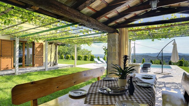 An outdoor patio with a wooden table set for dining, a bench, and a view of a swimming pool and mountains in the background. - Villa in Lupoglav With Pool Near the Sea (Lupoglav)