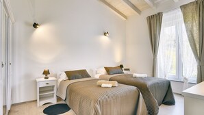 Une chambre avec deux lits, une table de chevet, une lampe, une fenêtre avec des rideaux et un plafond avec des poutres apparentes.