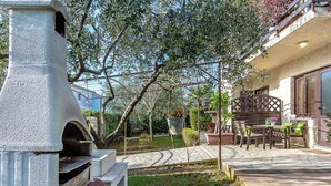 Un área de cocina exterior de piedra con una parrilla, rodeada por un jardín con plantas en macetas y un patio con mobiliario exterior.