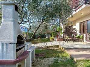 A stone-built outdoor kitchen area with a grill, surrounded by a garden with potted plants and a patio with outdoor furniture. - Inviting Apartment in Vinkuran near Sea (Medulin)