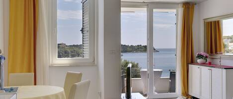 A room with a dining table, chairs, a red ottoman, a computer on a desk, and a view of the sea through large windows.