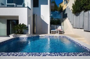 Ein moderner Außenpool mit Fliesenrand, eine kleine Sitzgelegenheit mit Stuhl und Tisch sowie eine Treppe, die zu einem Gebäude führt.