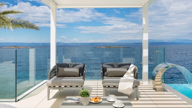 A balcony with two chairs, a table with a plant and pastries, and a glass railing overlooking the sea. - Beach Villa in Korcula with Pool, Sauna & Hot Tub (Blato)