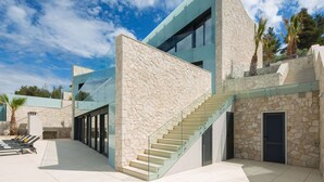 A modern building with a stone facade, a glass staircase, and a pool area with lounge chairs. - Beach Villa in Korcula with Pool, Sauna & Hot Tub (Blato)
