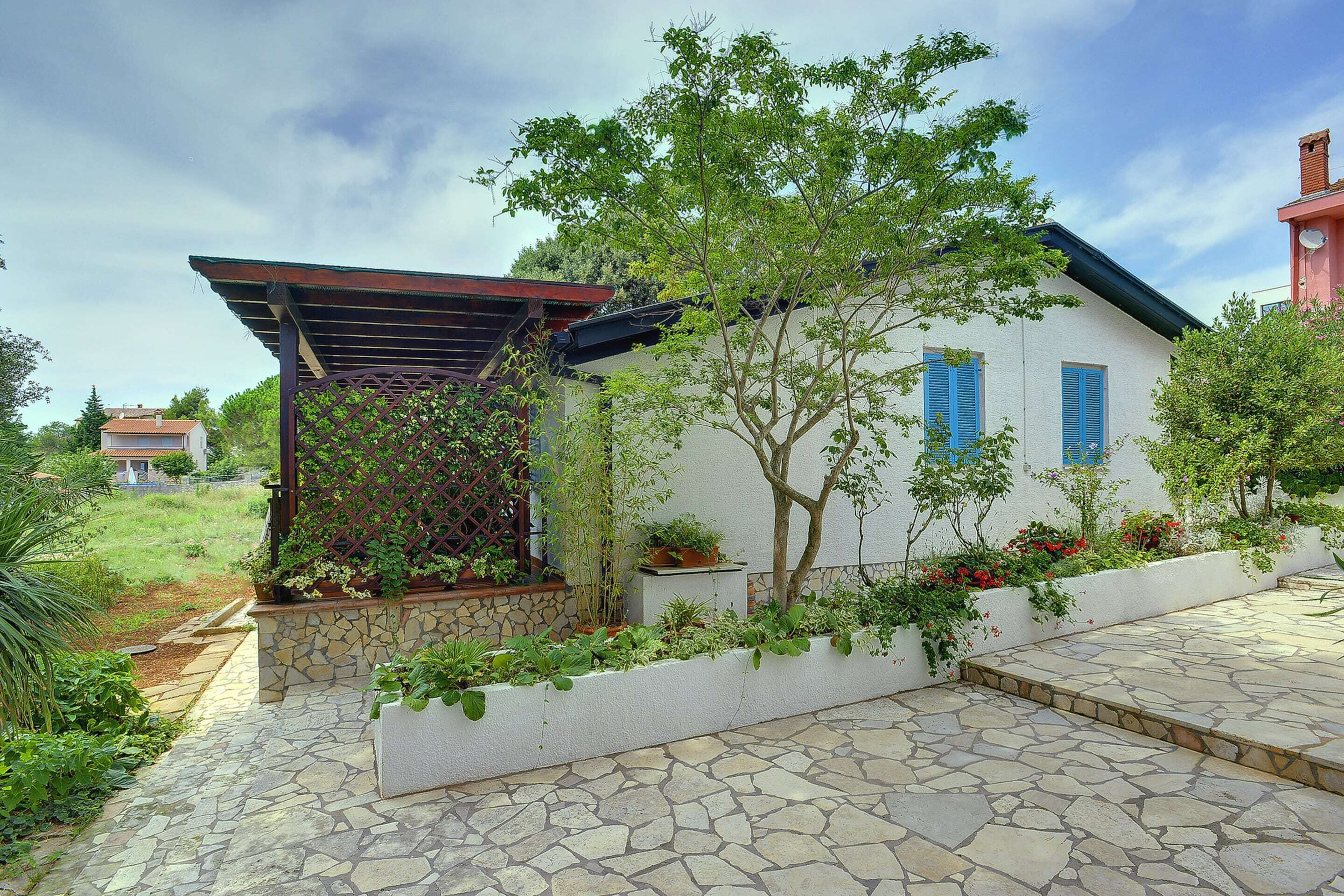 Ein Haus mit einer Steinterrasse, einer Pergola und einem Garten mit verschiedenen Pflanzen und Blumen.