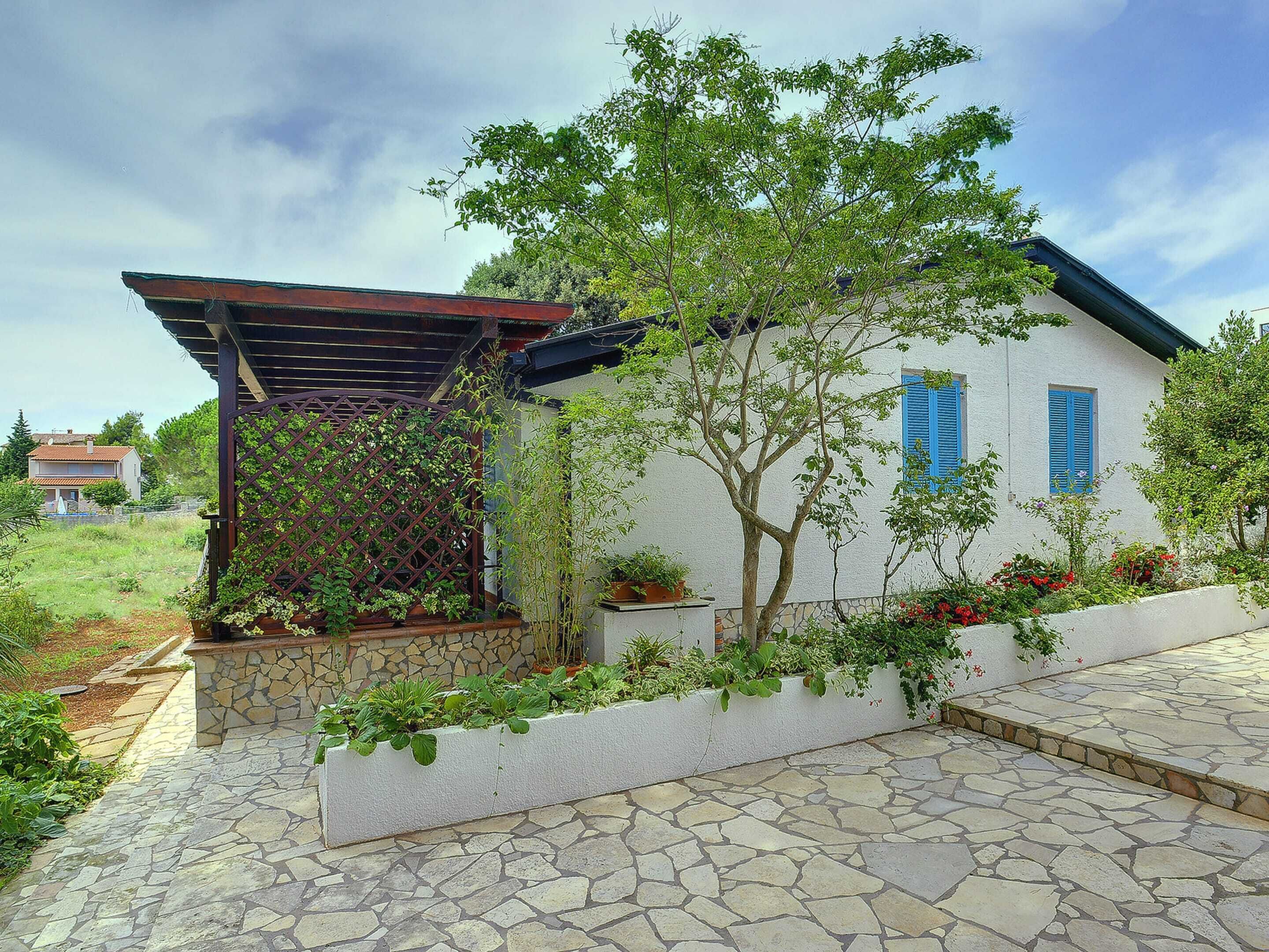 A house with a stone pathway leading to the entrance, a pergola with climbing plants, and a small garden area with potted plants and flowers.