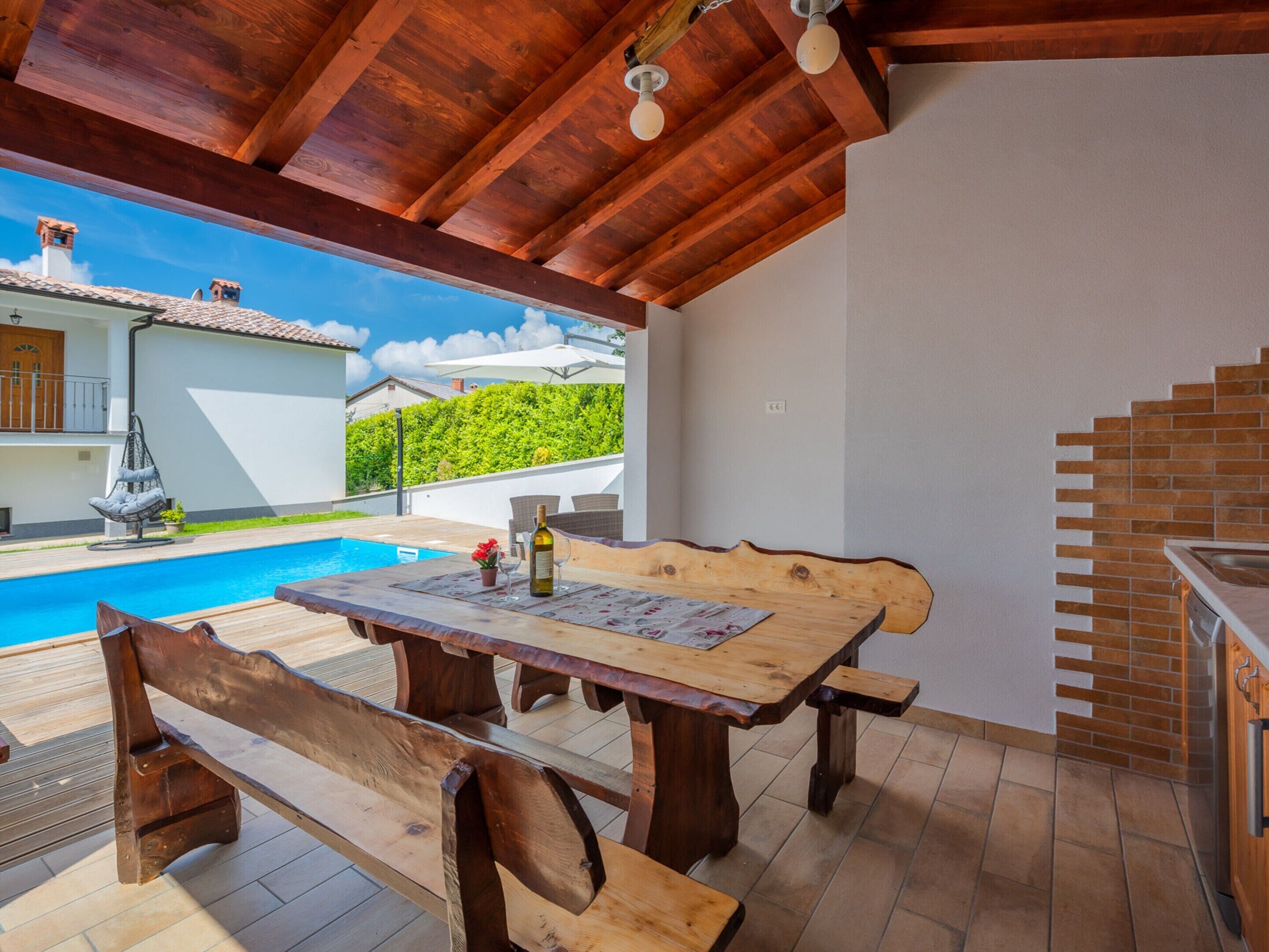 An outdoor patio with a wooden table and benches, a pool, and a view of a white house with a tiled roof.