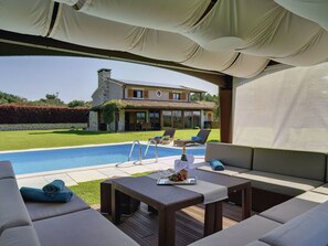 Ett utomhusområde med en pool, solstolar och ett bord, en täckt uteplats och ett hus i bakgrunden.