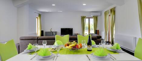 Ein Essbereich mit Tisch für vier Personen, umgeben von grünen Stühlen. Auf dem Tisch steht eine Obstschale und eine Weinflasche ist sichtbar. Der Raum hat ein modernes Design mit weißen Wänden und einem großen Fenster.