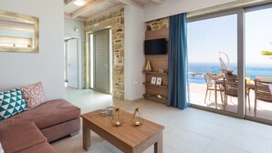 A modern living room with a wooden ceiling, a sofa with patterned cushions, a wooden coffee table with decorative items, a stone wall, and a large sliding glass door leading to an outdoor seating area with a view of the sea. - Scenic Villa With Private Pool (Kissamos)