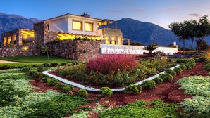 Une maison moderne aux murs en pierre, dotée d’un jardin bien entretenu, avec des montagnes en toile de fond.