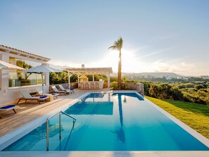 A modern outdoor pool area with a bar and lounge chairs, surrounded by a well-maintained lawn and a view of the city in the distance. - Beautiful Luxe Villa in Private Olive Grove, sea and Mountain View, Close to sea (Agios Nikolaos)