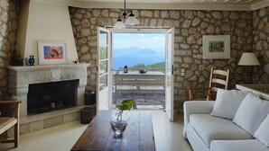 A cozy living room with a stone wall, a fireplace, a wooden table, and a white sofa. - Spacious Villa in Vasiliki with Swimming Pool & Hot Tub (Lefkada)