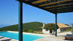 A pool area with stone flooring, lounge chairs, and a view of a hill and a building. - Spacious Villa in Vasiliki with Swimming Pool & Hot Tub (Lefkada)