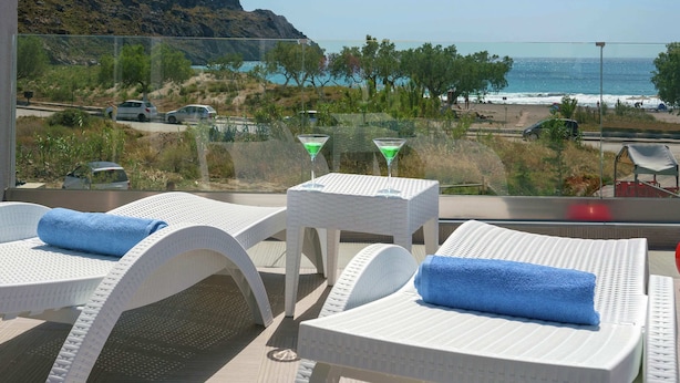 Ein Balkon mit weißen Korbstühlen und einem Glastisch, mit Blick auf einen Strand und ein Meer, im Hintergrund ein Berg.
