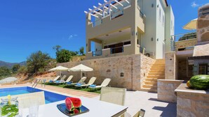 A poolside area with a table, chairs, and umbrellas. There is a stone wall and a building with balconies. - Splendid Villa in Achlades with Private Pool near Bali (Mylopotamos)