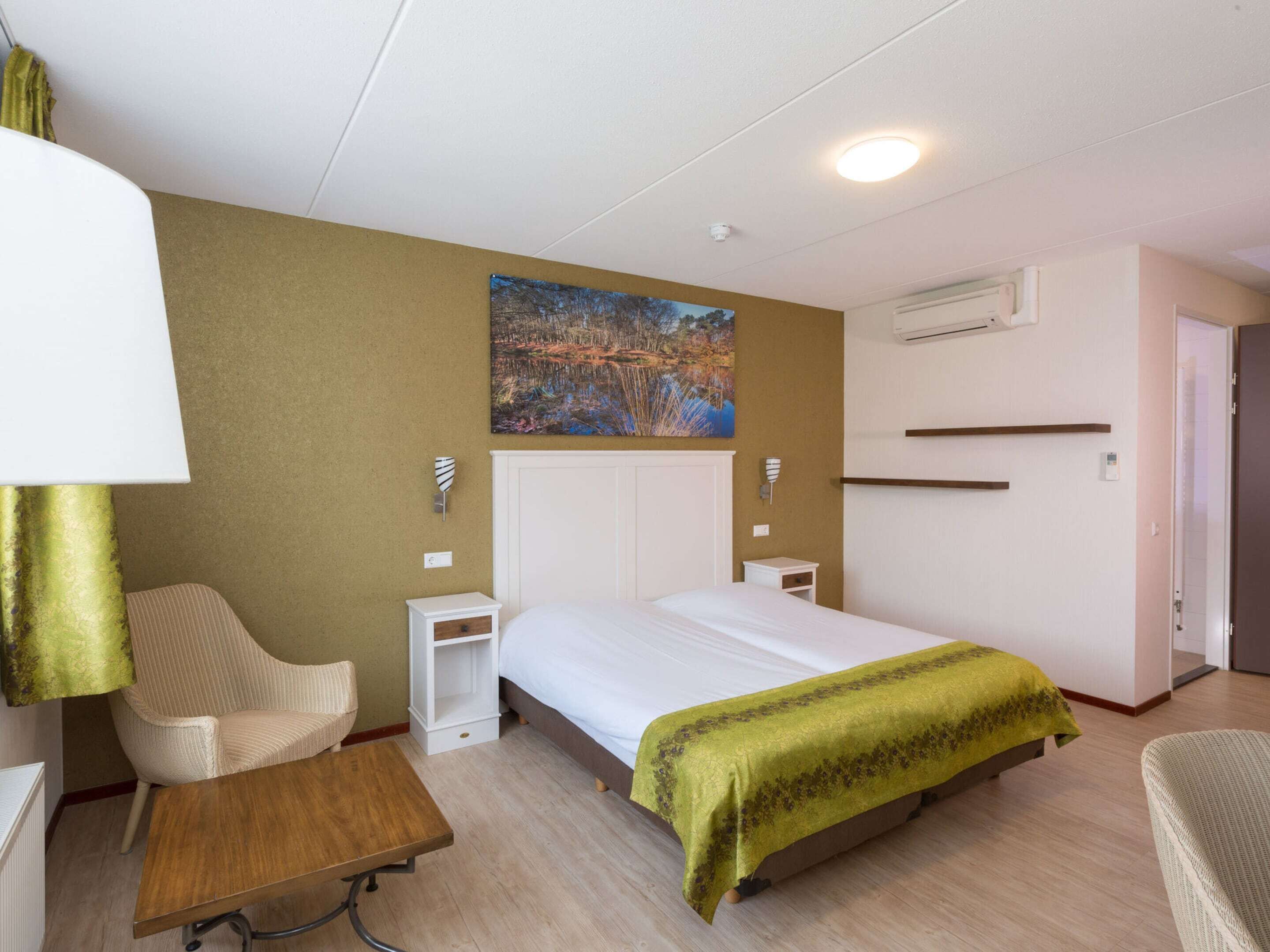 Una habitación de hotel con cama, una silla, una mesita, un equipo de aire acondicionado y un cuadro en la pared.