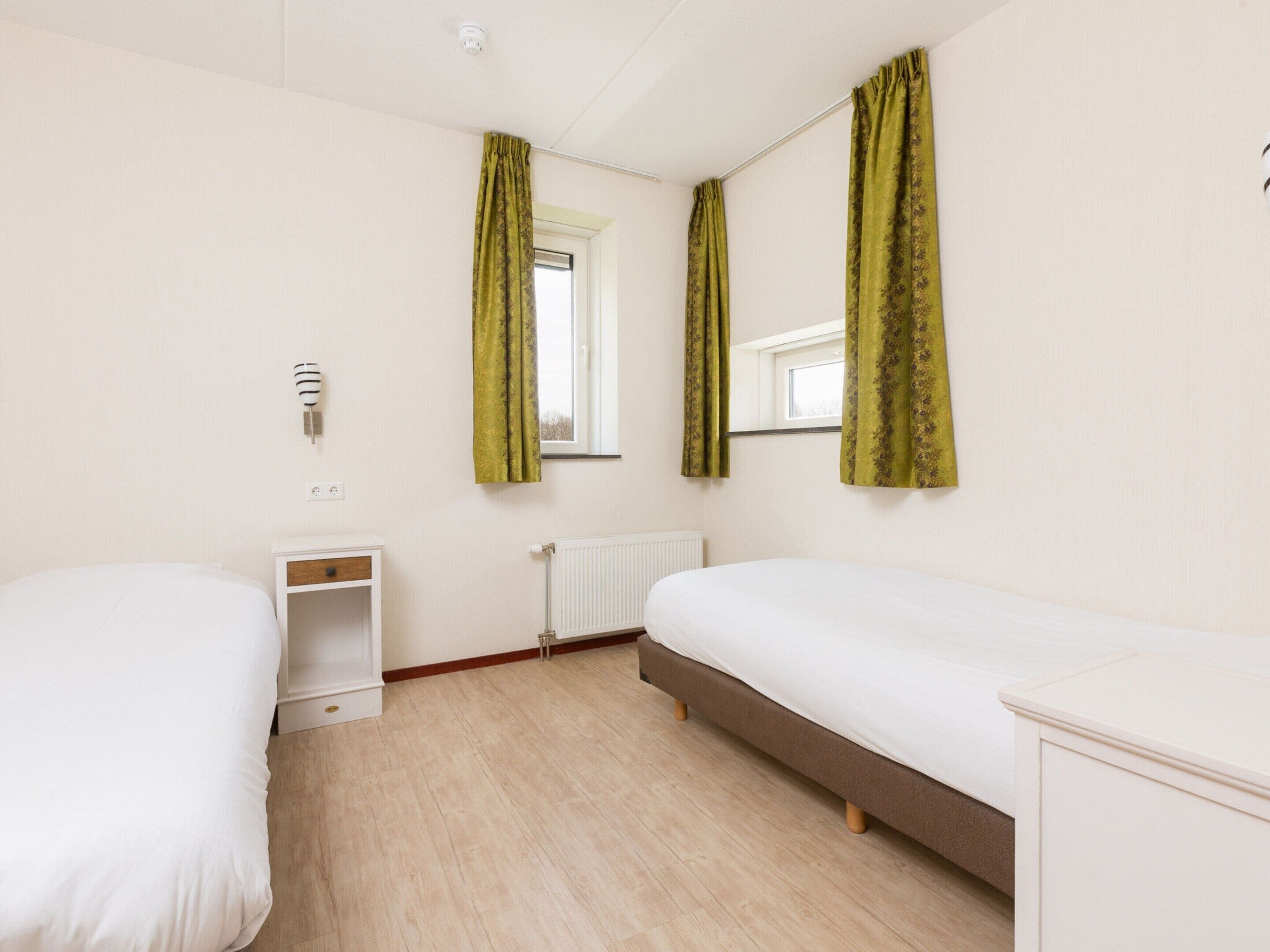 Une chambre avec deux lits simples, une table de chevet, un radiateur et une fenêtre avec des rideaux.