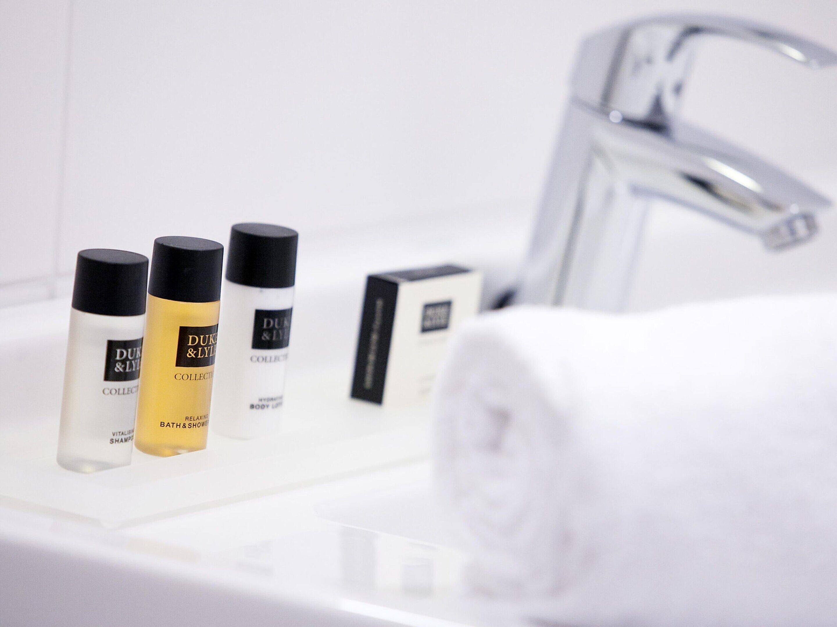 Une salle de bain avec un lavabo blanc, trois flacons de lotion pour le corps de la collection Duke & Lyall, et une serviette blanche enroulée.