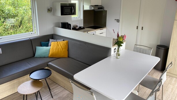 A compact kitchen with a microwave, stove, and sink. There is a small dining table with chairs, a sofa with cushions, and a window with a view of greenery. - Chalet in Friesland Near Nature Reserves (De Veenhoop)