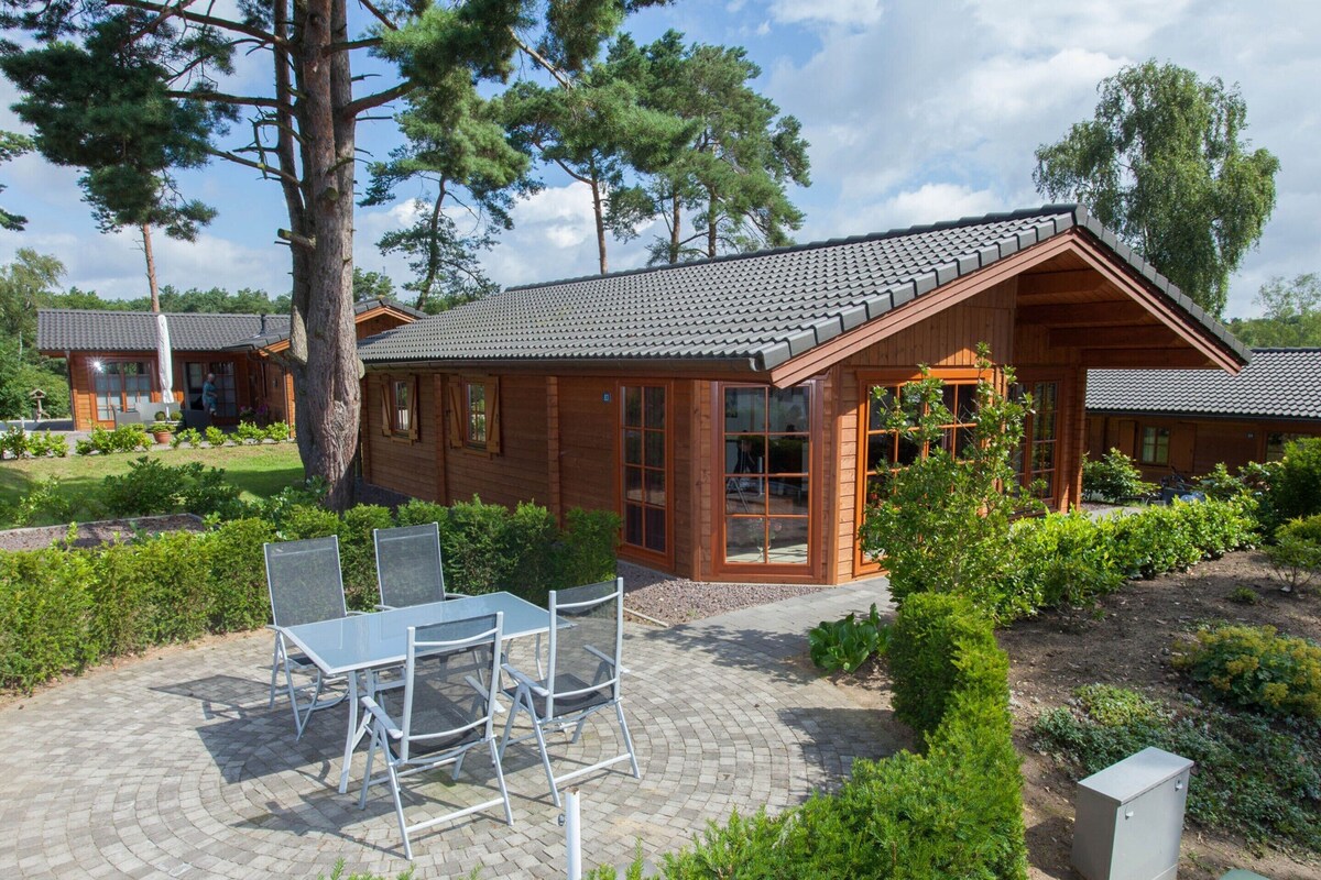 A wooden cabin with a tiled roof, a patio with a table and chairs, surrounded by greenery and trees.