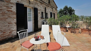 An outdoor patio with two white wicker chairs and a small round table, a brick building with a white door and black shutters, and potted plants. - Villa in Montemaggiore With Private Pool (Colli al Metauro)