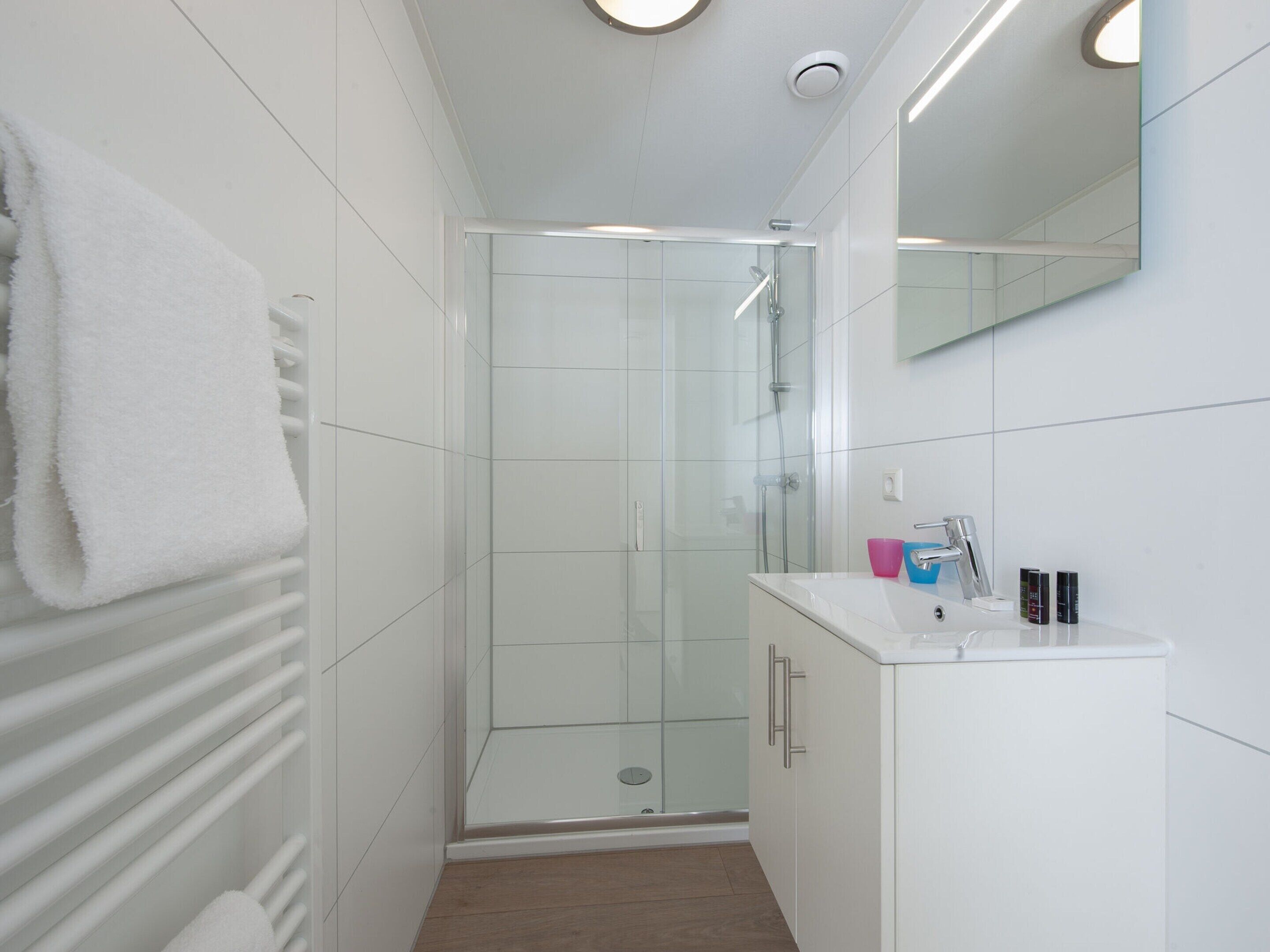 A modern bathroom with a glass shower enclosure, white tiled walls, a white vanity with a mirror, and a towel hanging on a radiator.
