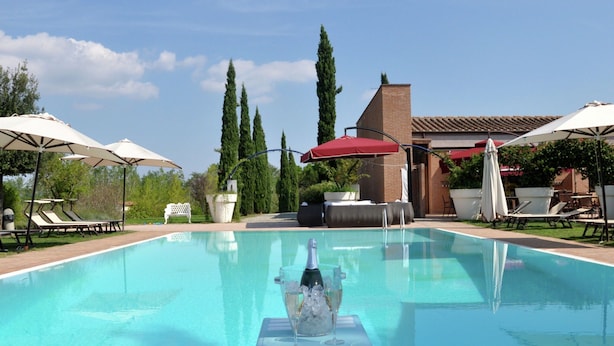 A swimming pool with lounge chairs and umbrellas, surrounded by tall trees and a building with a tiled roof. - Cozy Apartment With Terrace in Tuscany (Capannoli)