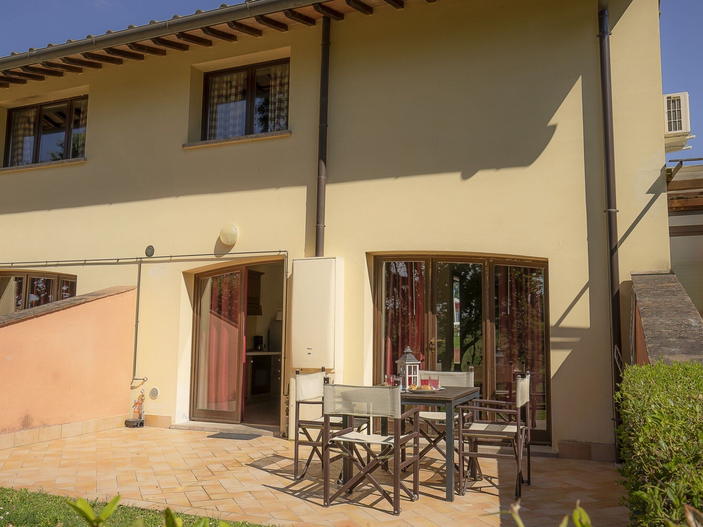 A two-story building with a patio area featuring a table and chairs, a small staircase, and a window with curtains.