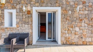 Un bâtiment en pierre doté d’une porte coulissante en verre qui donne sur une pièce où se trouve un lit. Il y a un banc devant la porte.