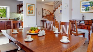 Uma sala de jantar ampla com uma mesa de madeira posta para quatro pessoas, rodeada por cadeiras. Há um prato de frutas e xícaras brancas sobre a mesa. Uma escadaria leva a um andar superior.