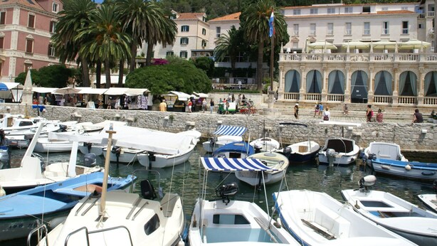 A marina with various boats docked, a historic building with arches, and a castle on a hill in the background.