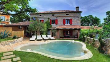 A stone house with a swimming pool, red shutters, and a garden with lounge chairs.