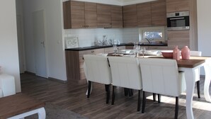 A modern kitchen with wooden cabinets, a dining area with white chairs, and a built-in oven. - Comfortable Chalet With Dishwasher (IJhorst)